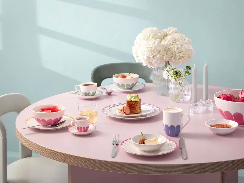 Ein runder Tisch mit Tellern, Tassen und Schüsseln voller Essen, dekoriert mit rosa Mustern. In der Mitte stehen eine Vase mit weißen Blumen und zwei unbeleuchtete Kerzen. Stühle umgeben den Tisch.