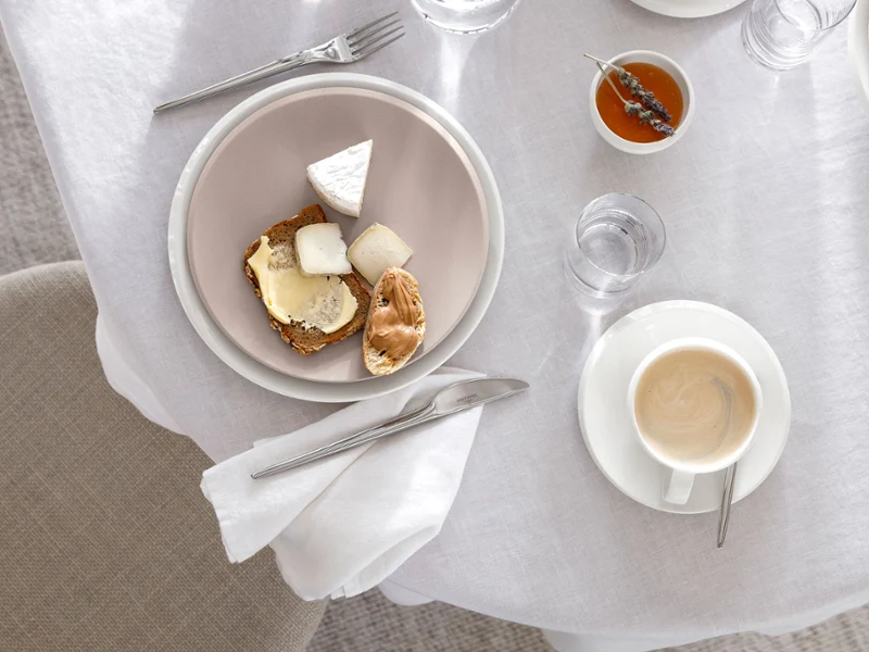 A set table with a cup of coffee, a NewMoon plate in beige with pastries, a small bowl of jam, and cutlery on a white tablecloth.