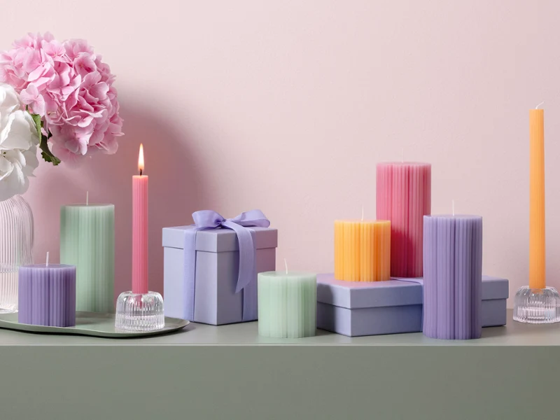 Velas Fleur en diferentes colores pastel y formas, una idea ideal para regalar Varias velas en colores pastel de Fleur y cajas decorativas para regalo sobre una superficie verde con flores rosas y blancas al fondo.