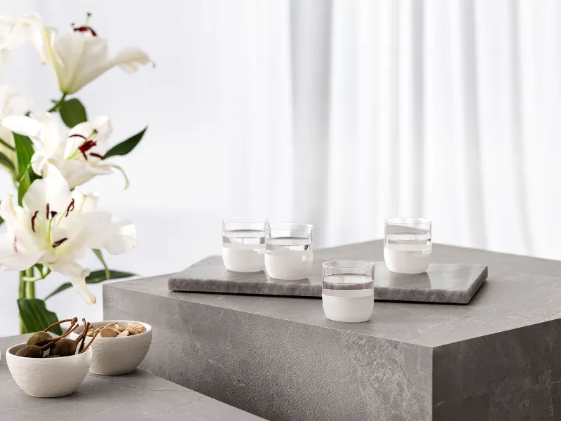 Four white Manufacture water glasses on a concrete table, together with white lilies in a vase and Manufacture bowls with snacks.