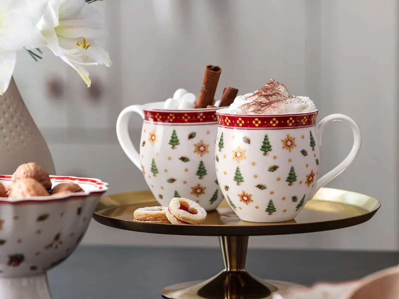 Dwie świąteczne filiżanki Toy's Delight z bitą śmietaną i laskami cynamonu na tacy, a obok nich herbatniki i wazon z kwiatami.