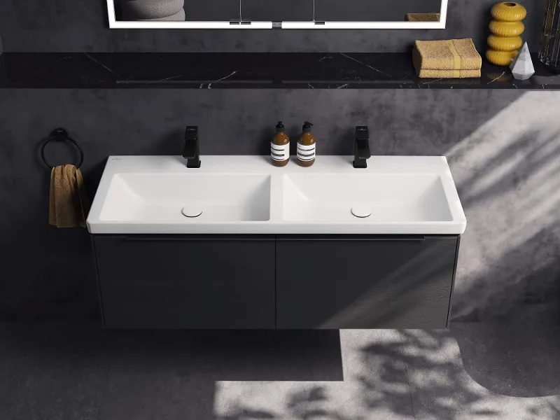 Badezimmer-Waschtisch im Allblack Stil Schwarzer 'Subway 3.0'-Badezimmerschrank mit weißen Doppelwaschbecken und schwarzen Armaturen, einem an der Wand montierten 'My View Now'-Spiegel von Villeroy & Boch und dekorativen Gegenständen wie Seifenspendern und gestapelten Handtüchern.