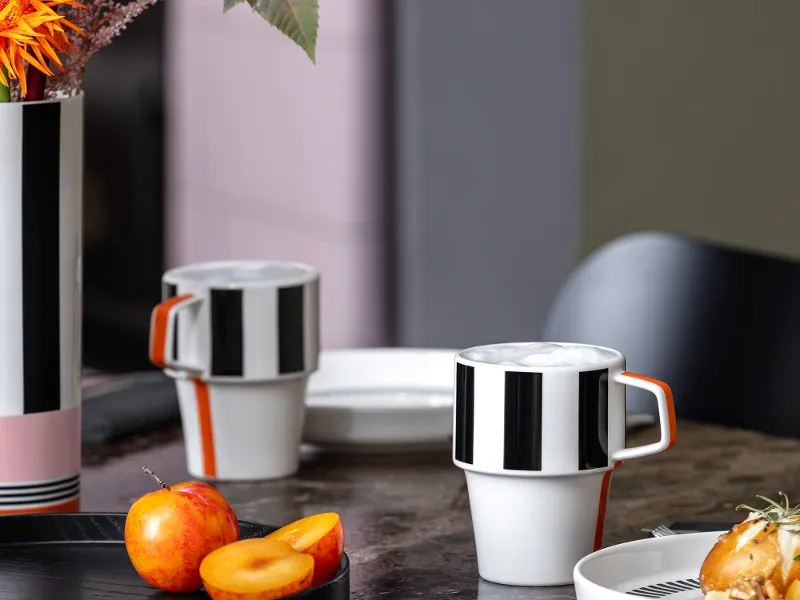 Modernes Tischgedeck mit gestreifter Memphis Vase, passenden Tassen, Obst und einem Brötchen auf dunkel marmoriertem Untergrund.