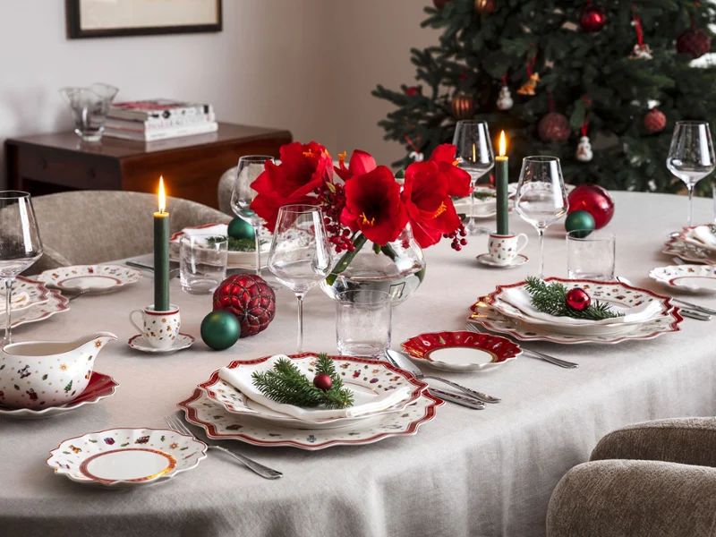 A dining table set for a festive meal A dining table set for a festive meal with red flowers, candles, and festive decorations next to a decorated Christmas tree.