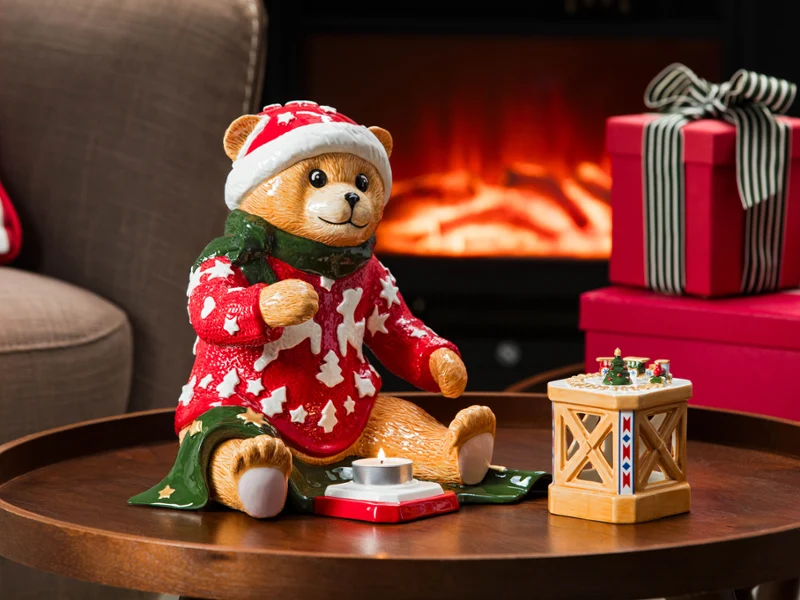 Ein Teddybär in festlicher Kleidung von Villeroy & Boch sitzt auf einem Tisch neben einer Laterne und verpackten Geschenken; ein Kamin ist im Hintergrund zu sehen.