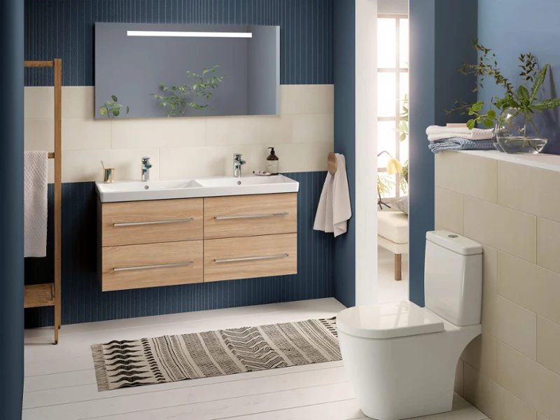 Baño moderno con lavabo doble, gran espejo, inodoro, paredes azules y beige, y luz natural desde una ventana.