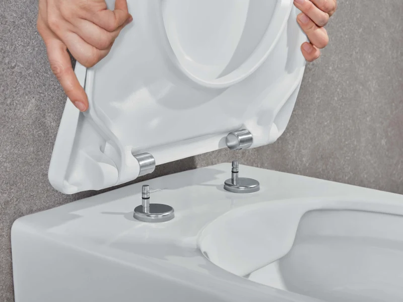 Mocowanie deski sedesowej Villeroy & Boch Osoba mocuje białą deskę sedesową do muszli klozetowej za pomocą dwóch metalowych śrub i zawiasów.