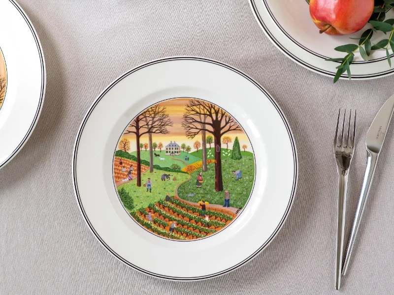 A decorative plate from the Design Naif collection by Villeroy & Boch depicts a rural scene with fields, trees, and people. The table setting includes a fork, a knife, and a pomegranate on the Naif design plate.