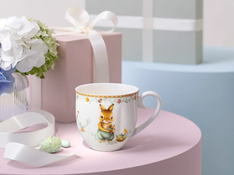 A decorated Annual easter Edition mug sits on a pink surface next to a white ribbon, pastel gift boxes, and a vase of flowers.