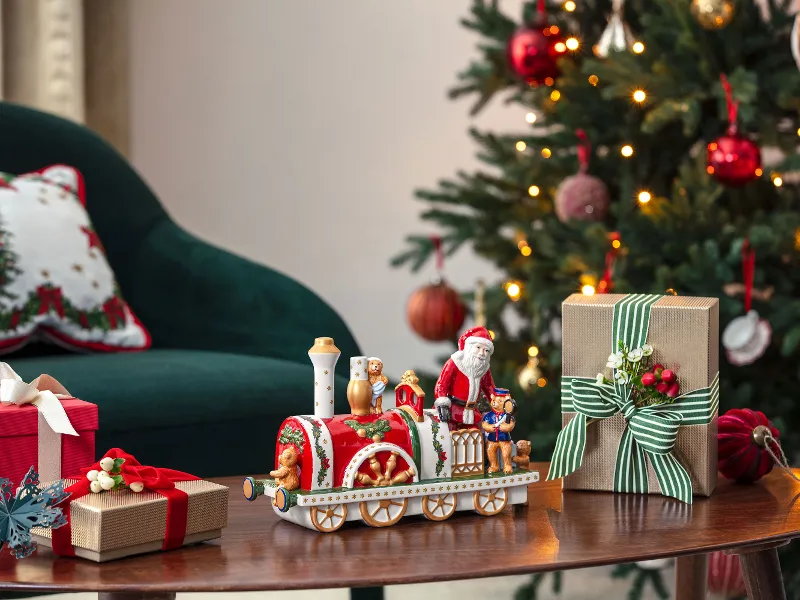  decorated Christmas tree, presents, and a Christmas Toys Memory locomotive on a wooden table next to a green chair.