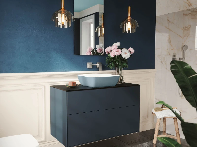 Bagno moderno con lavabo galleggiante blu navy, lavabo da appoggio, rubinetto a parete e grande specchio. Sopra sono appese due lampade a sospensione. Le pareti sono blu e bianche. Fiori rosa in un vaso e uno sgabello con un asciugamano.