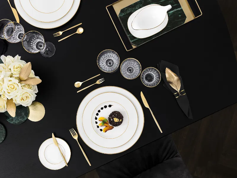 Elegant black table setting with gold Metrochic d'Or cutlery, white and gold-rimmed Anmut Gold plates, Grand Royal Gold glasses and a small bowl of food. A flower arrangement with white roses is also visible.