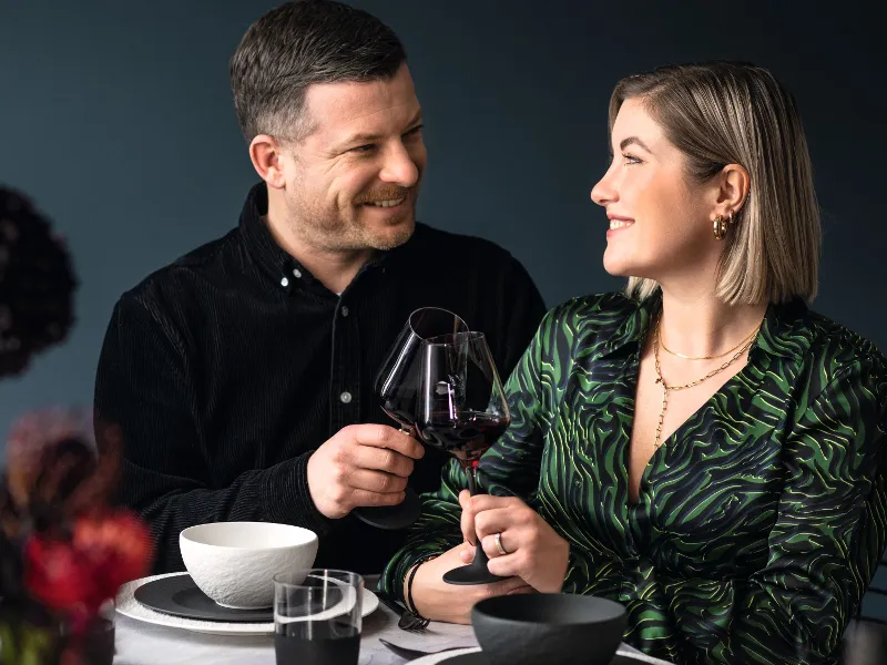 a man and woman having dinner toasting with red wine glasses and black and white dinnerware