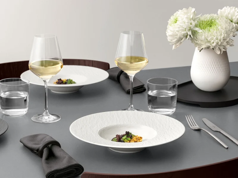 A formal table setting with a Manufacture pasta plate A formal table setting with a pasta plate ad food in it, glassware, and silver cutlery on a grey table.