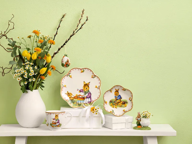 On a white shelf there is a vase with yellow flowers along with two decorative plates, a cup, a small figurine and a Villeroy & Boch "Annual Easter Edition" egg ornament. The wall in the background is painted light green.