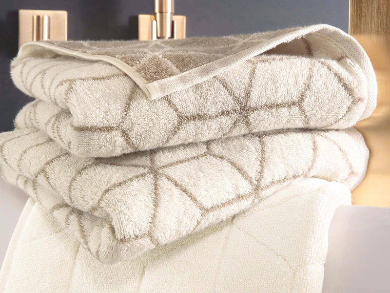 Elegancki wystrój łazienki z beżowymi ręcznikami Carré i złotym kranem. Stos złożonych beżowych ręczników Carré z geometrycznym wzorem marki Villeroy & Boch, umieszczonych obok złotego kranu.