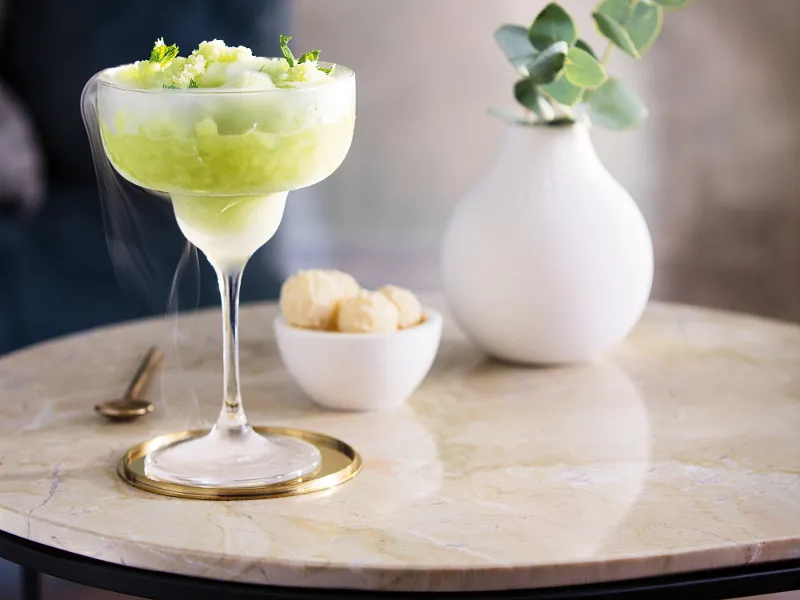 A chilled cocktail garnished with lime zest, served in a Purismo cocktail glass on a marble table, accompanied by a small bowl of snacks and a white vase with green plants.