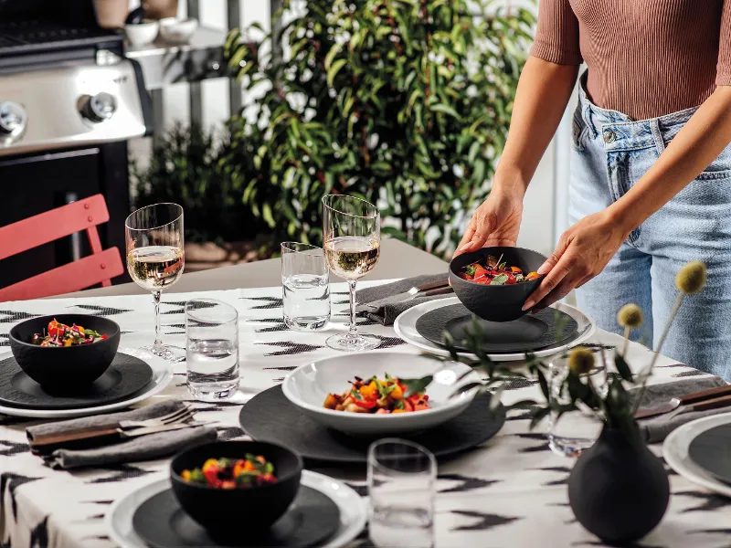 A woman setting a table with NewMoon and Manufacture plates and bowls for a barbecue evening outdoors.