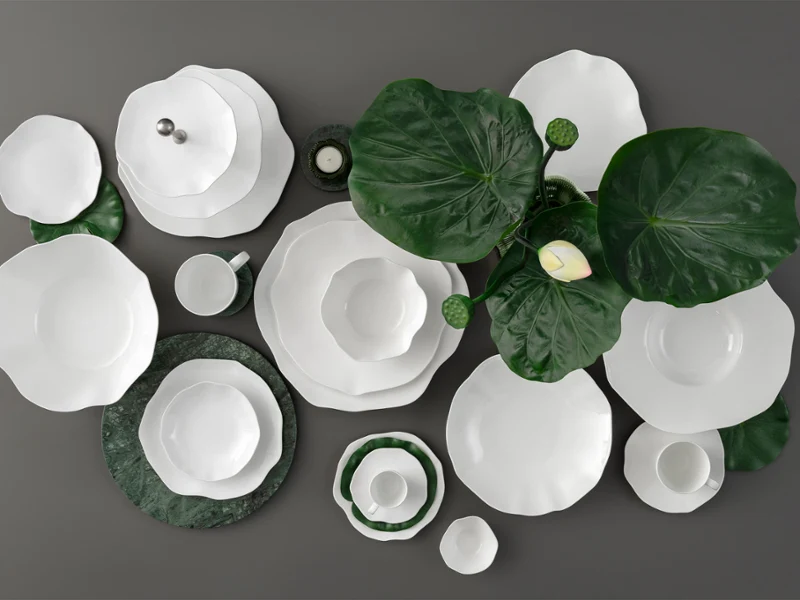 Weiße Geschirrteile der Kollektion Pura auf einer dunklen Fläche stehen. Weiße Geschirrteile der Kollektion Pura auf einer dunklen Fläche mit großen grünen Blättern und einer einzelnen weißen Blume als Dekoration.