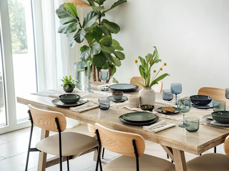 The dining table set with varios items from the Lave collection by Villeroy & Boch in green and grey, accentuated by blue glasses and napkins. A vase of yellow flowers and a small potted plant provide decoration, while a large plant stands by the window.