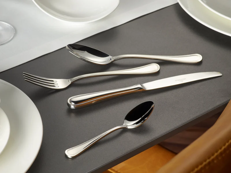 The Merlemont flatware set from Villeroy & Boch, consisting of a fork, knife, tablespoon, and teaspoon, is carefully arranged next to white plates and a bowl on a dark table.