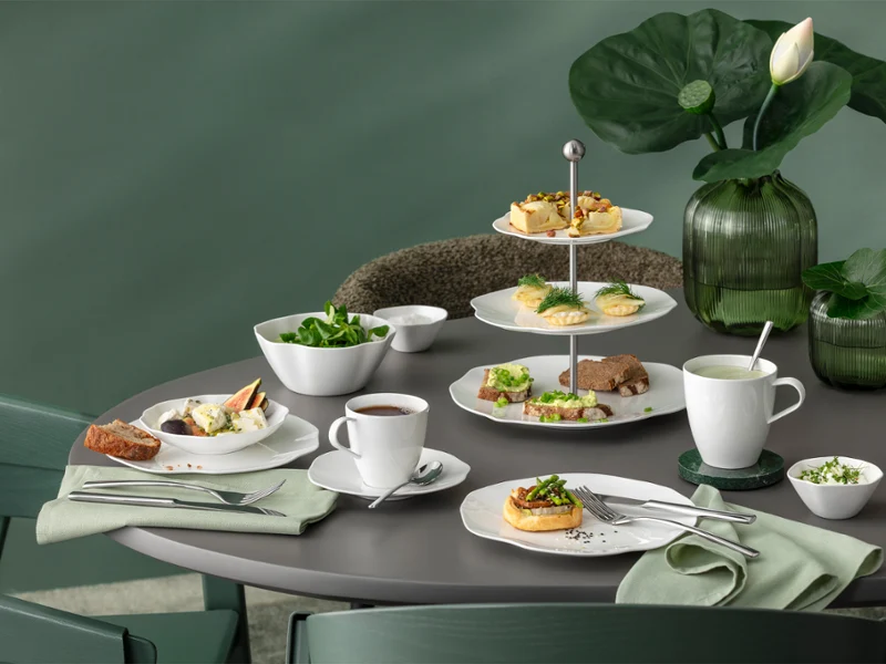 A round table with white tableware from the Pura collection against a green background A round table with white tableware from the Pura collection, various baked goods, salad, tea and green leaf decorations against a green background.