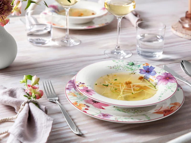 A table setting featuring floral-patterned "Mariefleur" plates by Villeroy & Boch, a soup bowl, cutlery (fork, spoon, knife), a napkin, two wine glasses, two water glasses, and a vase with flowers.