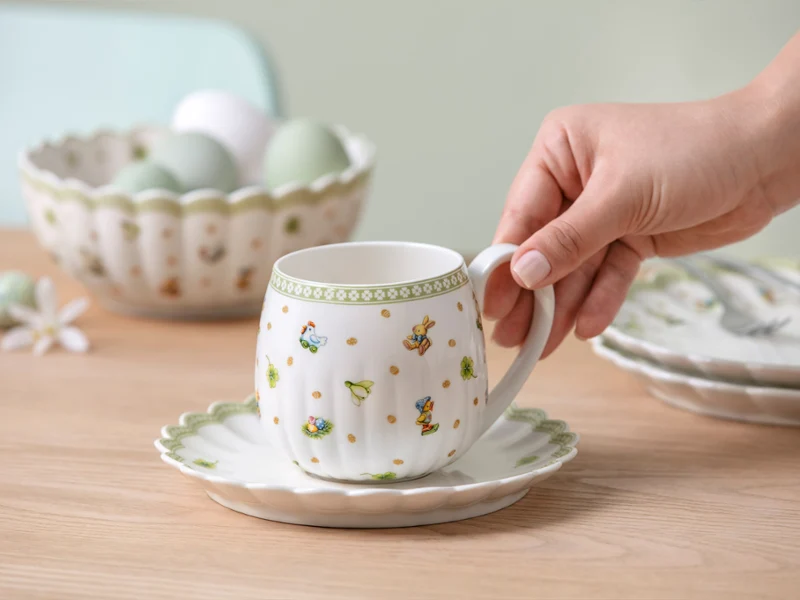 A hand holds a Easter Delight teacup on a saucer A hand holds a Easter Delight teacup on a saucer, with a bowl of eggs and plates in the background on a wooden table.