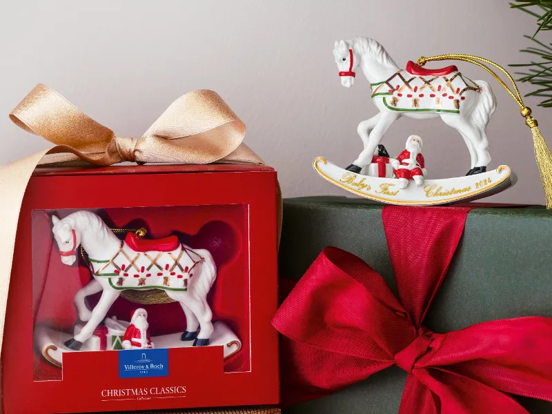 A festive presentation of two gift boxes: one with a Villeroy & Boch ornament with a rocking horse motif and another wrapped in green paper with a red bow. The rocking horse ornament from Villeroy & Boch, decorated with Christmas ornaments, can be seen above.