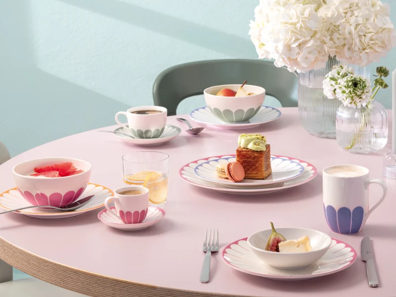A round table with pastel-coloured plates, cups and bowls filled with desserts and fruit, as well as a vase with white flowers.