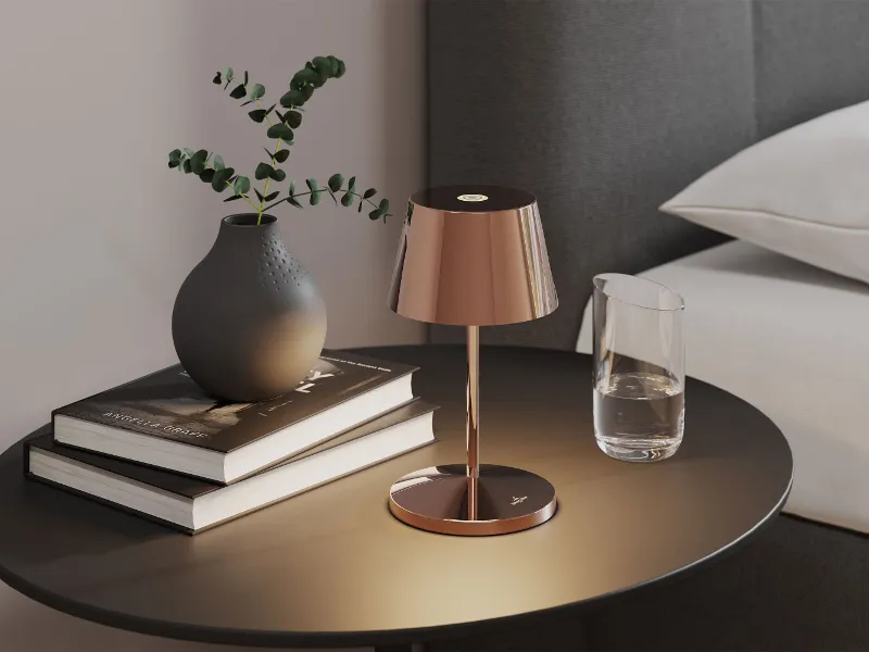  Table de chevet ronde avec une lampe dorée, deux livres, un vase sombre avec de la verdure et un verre d'eau à côté d'un lit. 
