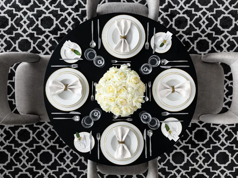 A round table with elegant white crockery from the Villeroy & Boch Anmut collection, napkins and Metrochic d'Or cutlery. In the middle is a centrepiece of white roses. The floor has a geometric black and white pattern.