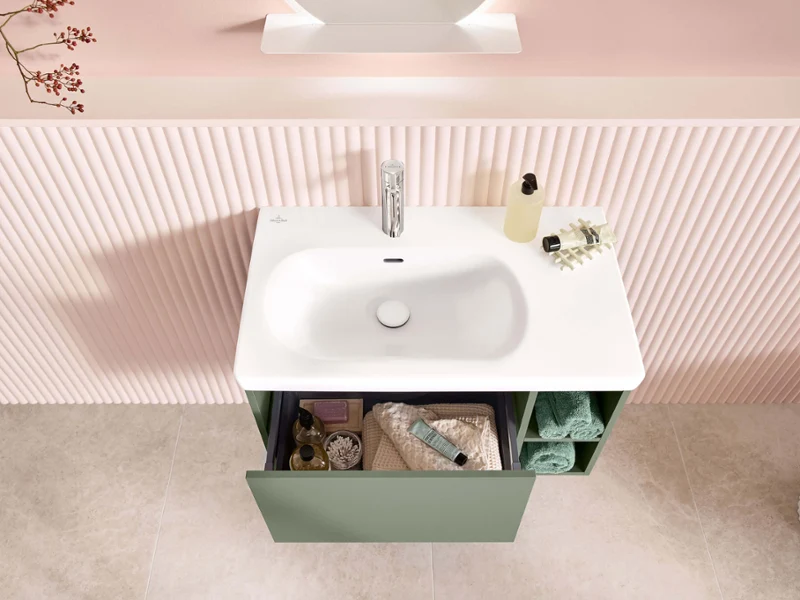 Trendy en stijlvol ingerichte Skyla-wasplaats in pasteltinten met binneninrichting De combinatie van wastafel, onderkast en chromen kraan uit de Skyla-badkamercollectie wordt van bovenaf getoond. De zachte, afgeronde vorm van de wastafel en de geopende lade van de kast zijn zichtbaar.