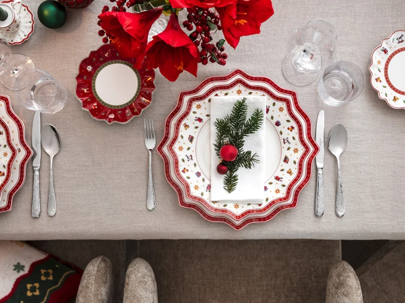 Toy's Delight crockery on a festively laid table Top view of an elegant festive table with the Villeroy & Boch Toy's Delight collection, complete with red and white plates, silver cutlery and a festive napkin decorated with pine wood and ornaments.