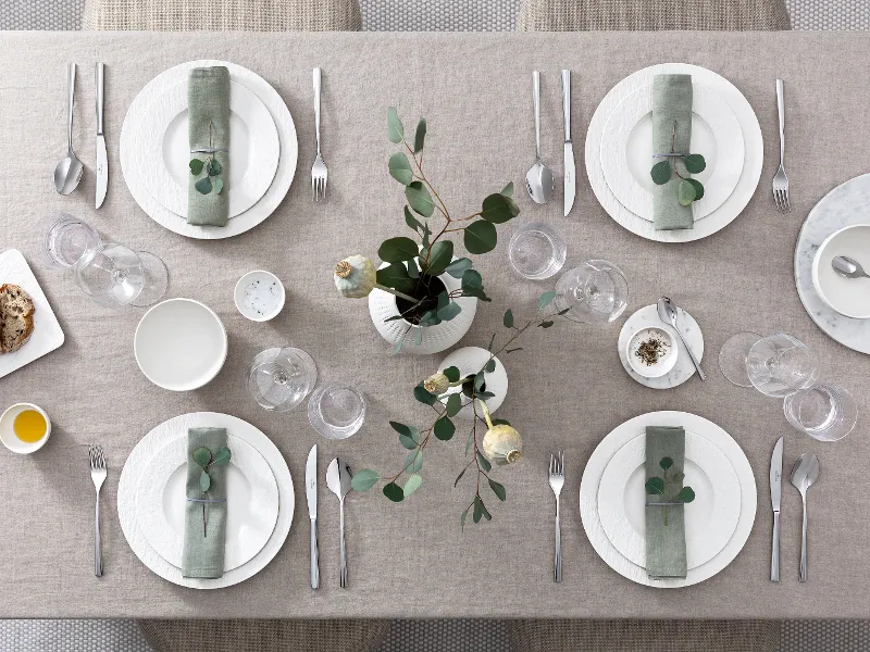 Modern dining table for six people with white Manufacture plates, cutlery and green floral decoration, seen from above.