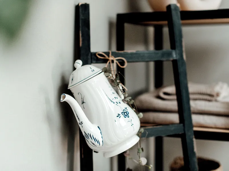 A white and blue Old Luxembourg teapot with a floral pattern hanging next to folded towels on a black wooden ladder.