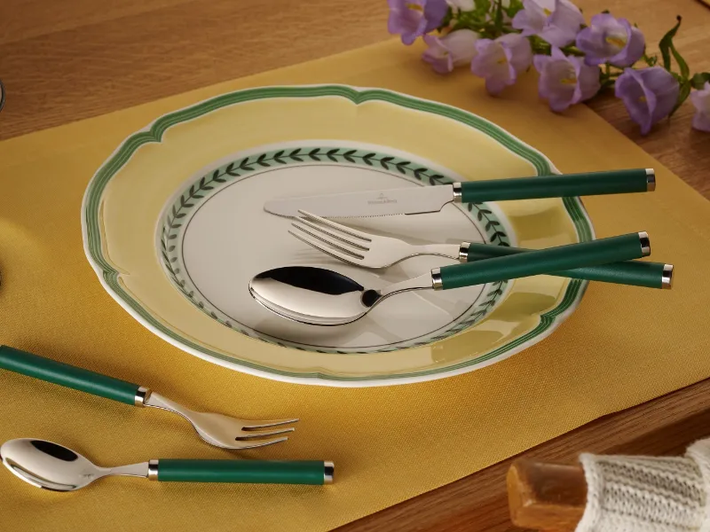 A dining room set is furnished with the Play! collection from Villeroy & Boch and features a decorative plate, flatware with a green handle, and a yellow placemat on a wooden table with purple flowers in the background.