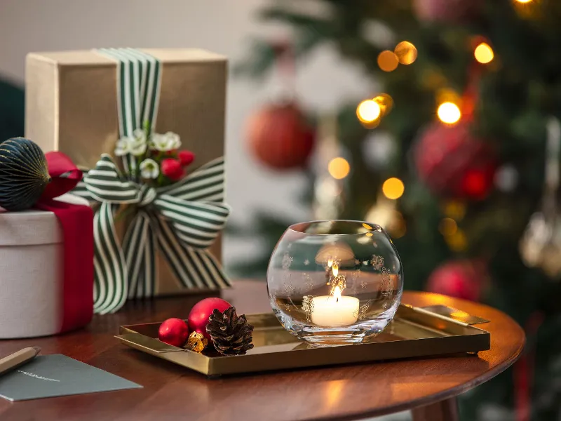 A burning candle in a Toy's Delight tea light holder stands on a tray next to wrapped presents, with a decorated Christmas tree in the background.