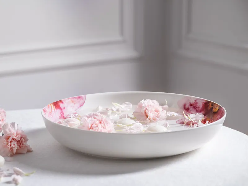 A Rose Garden bowl filled with water and floating pink flowers, surrounded by more petals on a white surface.