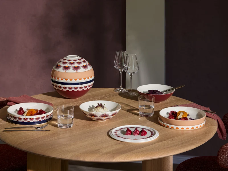 La Boule Kyoto tablesetting with food Ein runder Holztisch, gedeckt mit gemustertem Geschirr aus der La Boule Kyoto, Gläsern und einer Schüssel mit Deckel, bereit für ein Essen für drei Personen.