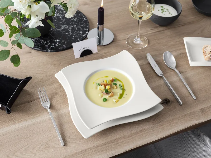 Eine Schüssel cremige Suppe mit Garnitur auf einem gedeckten Tisch mit Besteck, Wein, Blumen und einem kleinen Teller Brot.