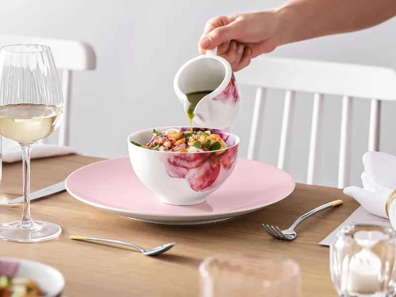 A person pouring dressing over a salad in a Rose Garden bowl at an elegantly laid dining table with Rose Garden crockery.