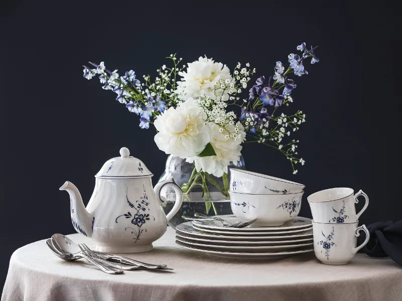 An Old Luxembourg porcelain set with a blue and white floral pattern, consisting of a teapot, cups and plates on a round table, with a vase of white and purple flowers in the background.