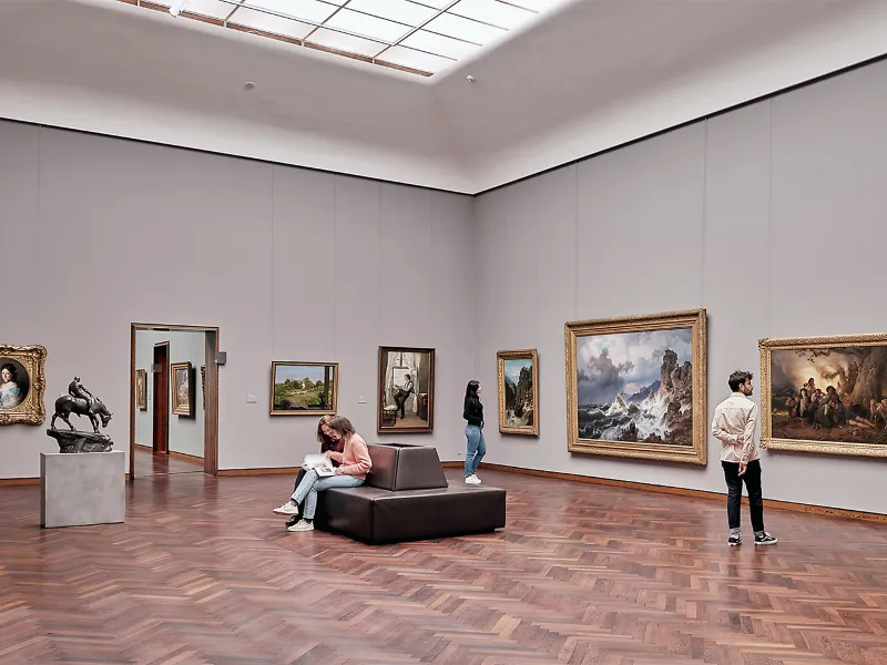 Four people are looking at paintings and a sculpture in a spacious art gallery with wooden floors and high ceilings.