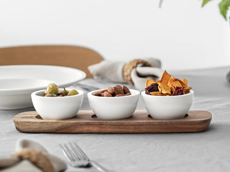 Three Artesano dip bowls with various snacks on a wooden serving tray, arranged on a table with elegant place settings and a stem with leaves.