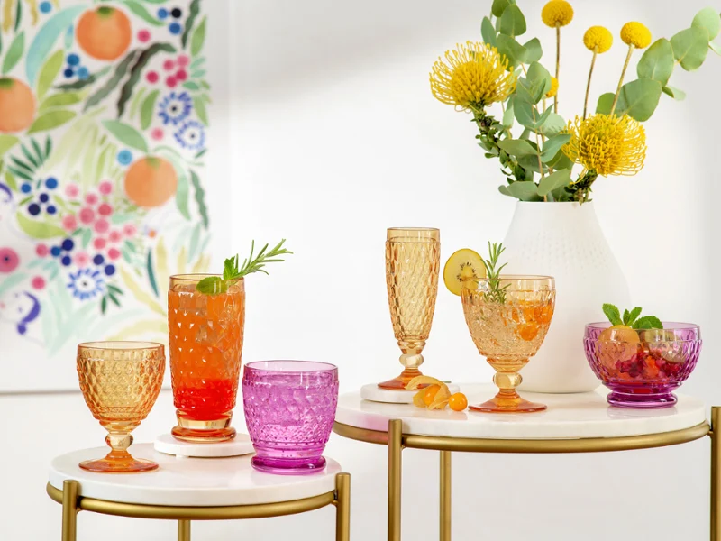 Colourful Boston glasses on tables with various drinks Colourful glasses on tables with various drinks, garnished with herbs and fruit slices. Yellow flowers and green plants in a vase. Wall art with floral and fruit patterns in the background.