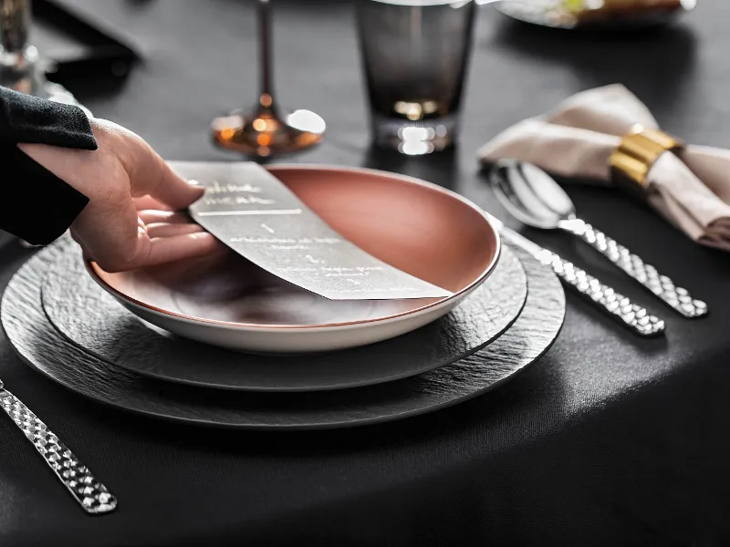 A person placing a menu card on an elegant table with a black tablecloth, cutlery and glasses together with copper-coloured and black Manufacture crockery.