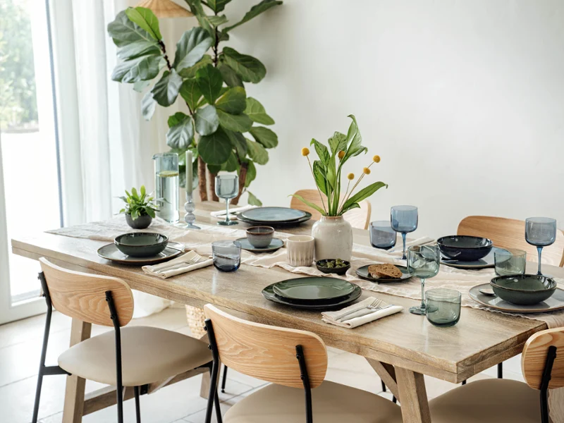 A dining table with green plates, beige napkins, blue glasses and small plants. Light-coloured wooden chairs surround the table. A large plant stands near a window.