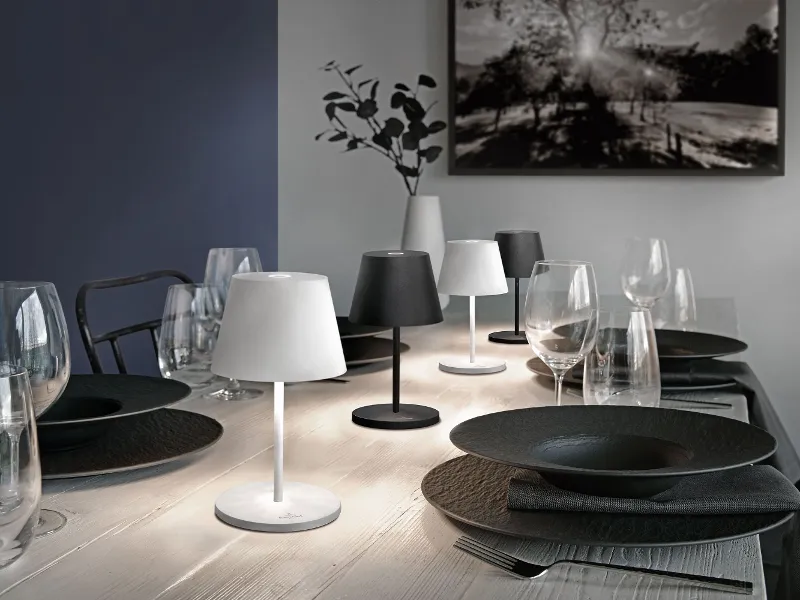 Ein moderner Esstisch mit schwarzen Manufacture Tellern, Gläsern und kleinen Tischlampen, im Hintergrund Kunstwerke und eine Vase.