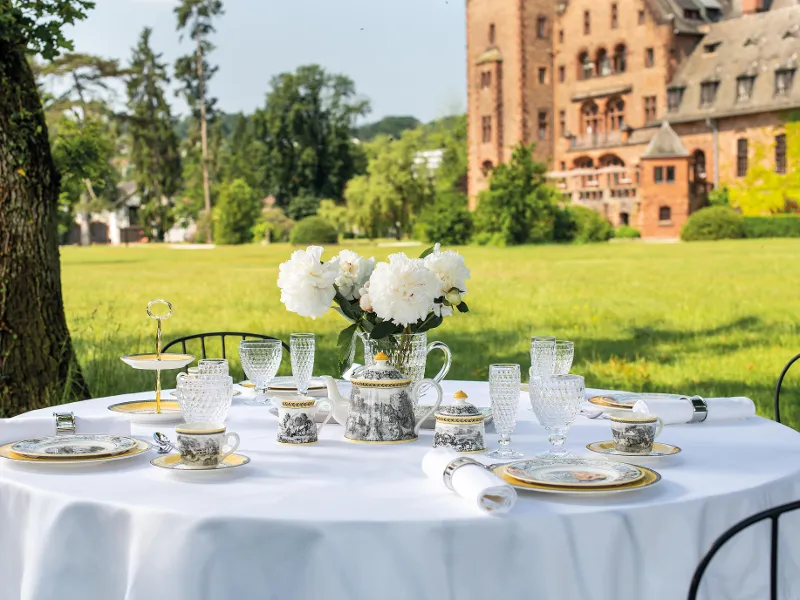 Elegant outdoor dining with fine Audun place settings and Boston glasses on a white tablecloth in front of a historic castle on a sunny day.